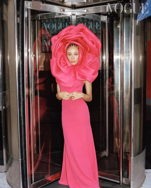 Woman in a pink dress with a large pink flower headpiece, standing in front of revolving doors with Vogue magazine covers visible.
