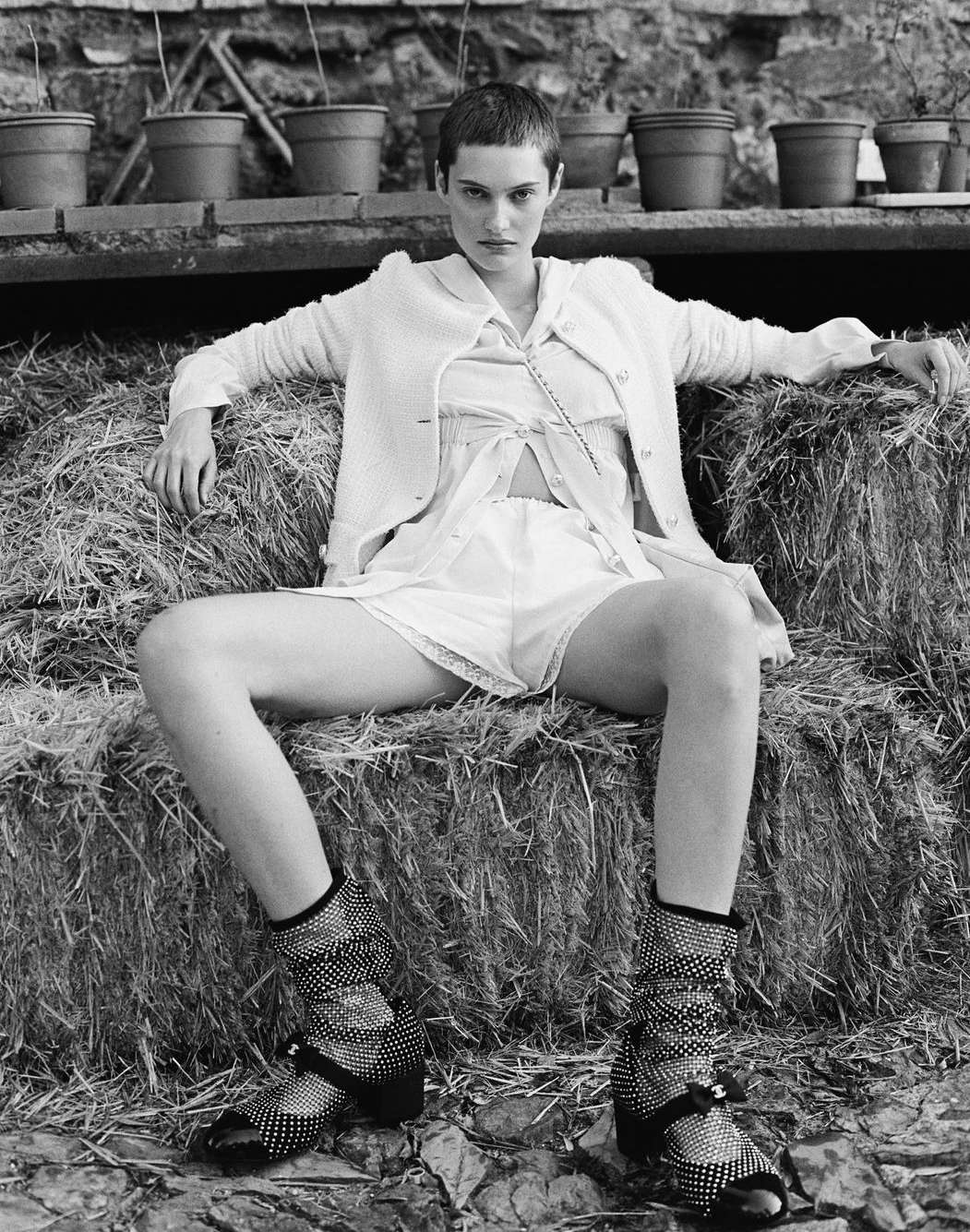 Monochrome shot of a fair-skinned young adult woman with short hair sitting on a hay bale wearing a short dress, cardigan, and polka-dot shoes.