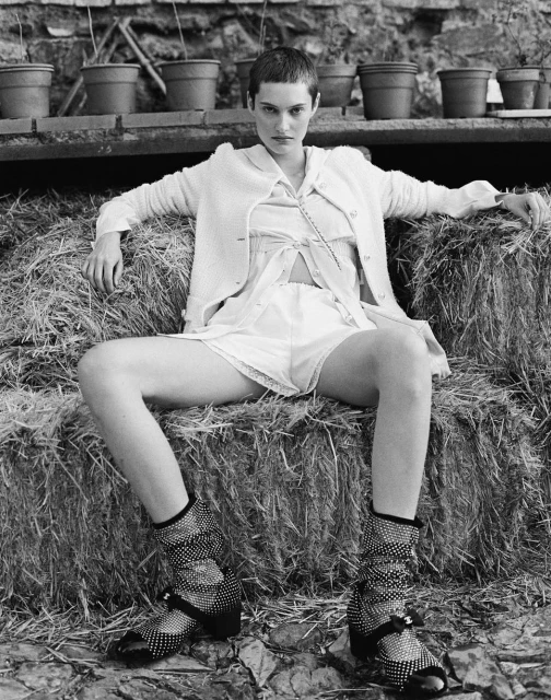 Monochrome shot of a fair-skinned young adult woman with short hair sitting on a hay bale wearing a short dress, cardigan, and polka-dot shoes.