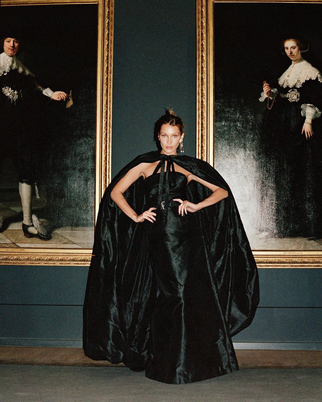 Bella Hadid poses in a black dress and cape at a museum, standing in front of two framed paintings with an elegant backdrop.