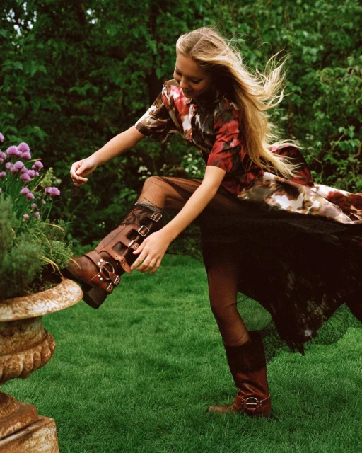 Woman in patterned top and tulle skirt stepping on stone planter with brown leather buckle boots in green grassy outdoor setting.