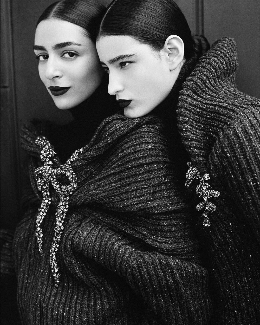 Black-and-white fashion shot of two women in dark turtlenecks and knitwear adorned with decorative brooches, showcasing dramatic dark lipstick.