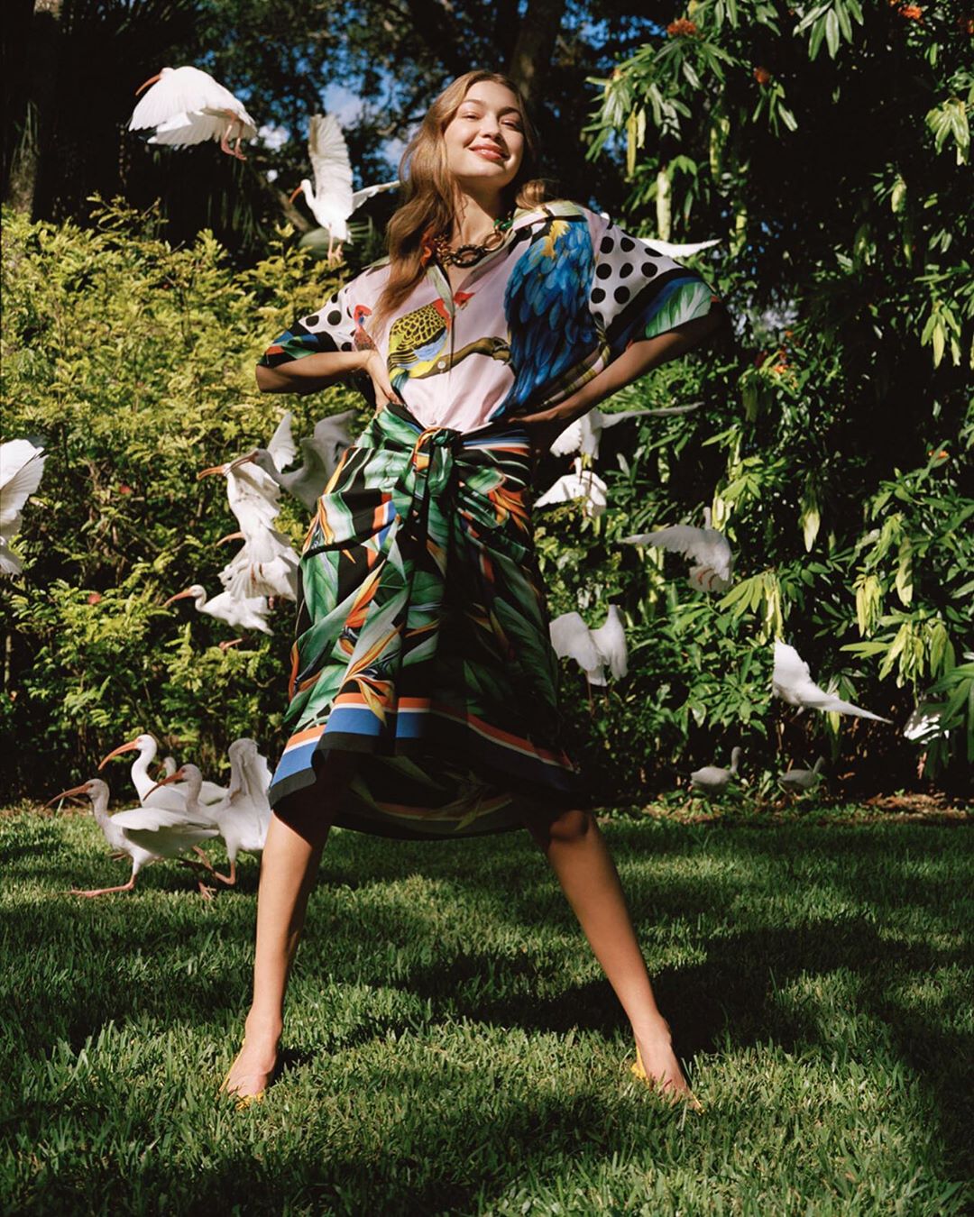 Young woman in patterned top and wrap skirt poses in a yard, encircled by white birds, lush greenery, sunny daylight and blue sky.