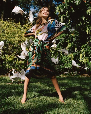 Young woman in patterned top and wrap skirt poses in a yard, encircled by white birds, lush greenery, sunny daylight and blue sky.