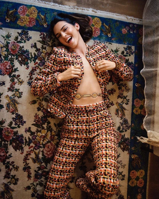 A woman with brown hair lies on a floral rug, wearing a tweed blazer and pants with a gold chain belt, smiling with an open mouth.