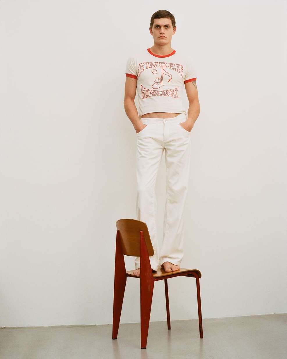 Full-length studio shot of a slim adult male standing barefoot on a wooden chair wearing a graphic tee and white pants.
