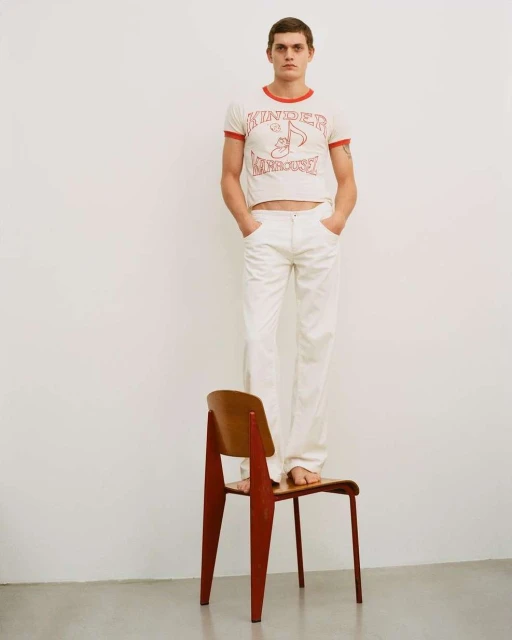 Full-length studio shot of a slim adult male standing barefoot on a wooden chair wearing a graphic tee and white pants.