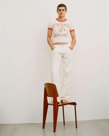 Full-length studio shot of a slim adult male standing barefoot on a wooden chair wearing a graphic tee and white pants.