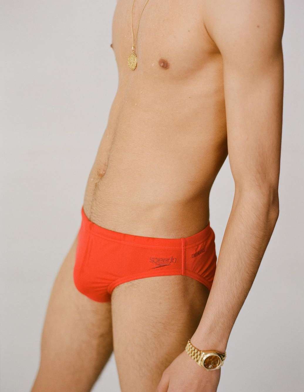 Adult man in red Speedo swimwear, side view. Gold watch and necklace. Light grey background. Fashion, fitness, and swimwear.