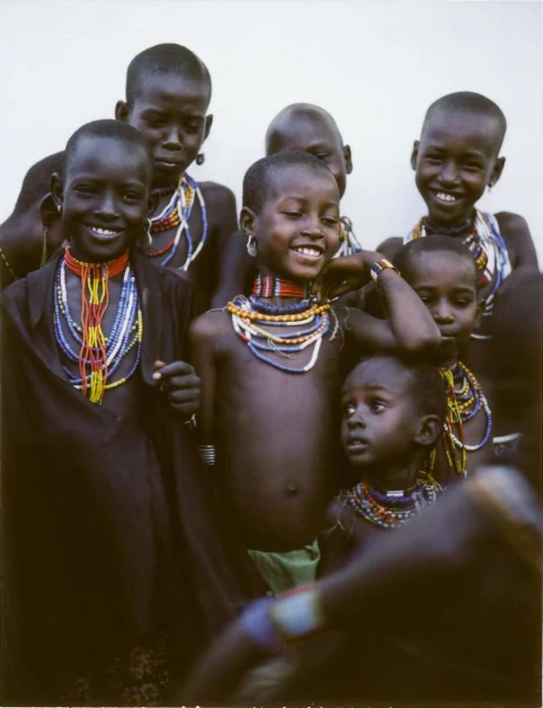 Group of smiling African children in traditional beaded necklaces and dark clothing against a plain background, medium shot.
