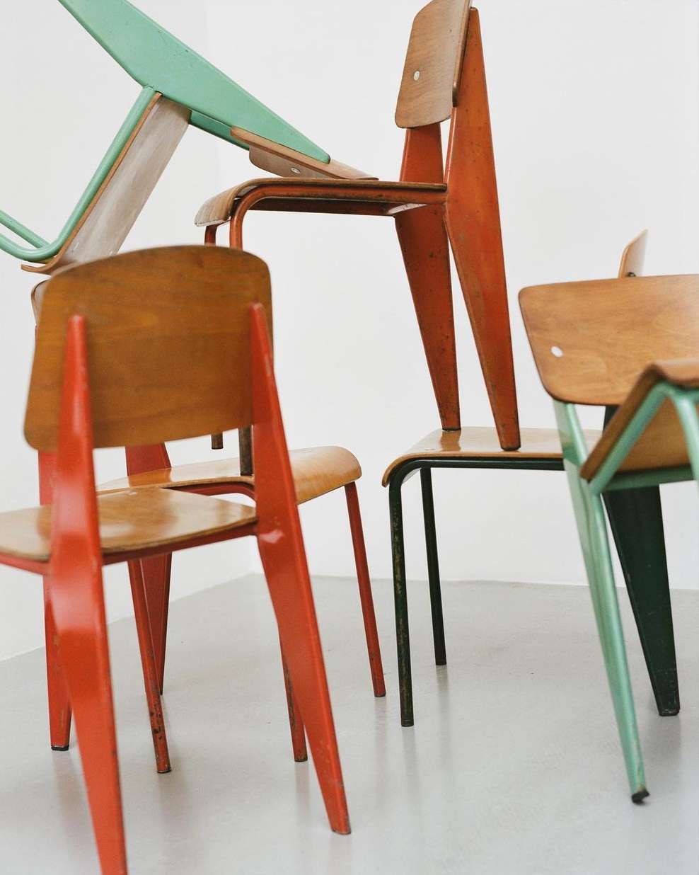 Stack of colorful vintage wooden chairs with metal frames, indoor setting on a gray floor against a white wall.