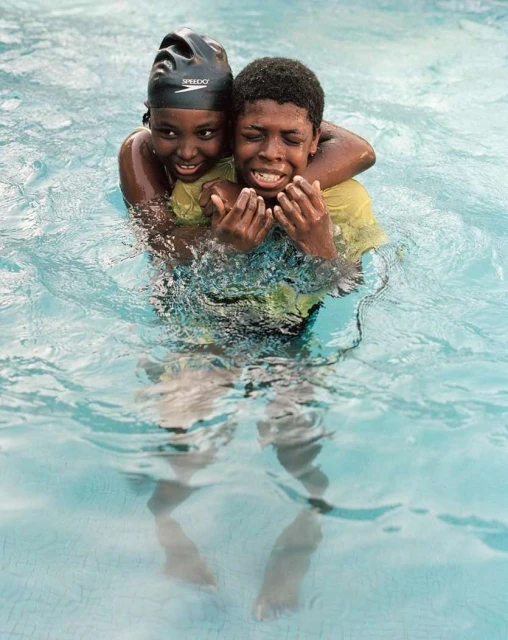 A smiling young woman in a swim cap embraces a distressed young man in a pool, both partially submerged in clear blue water.