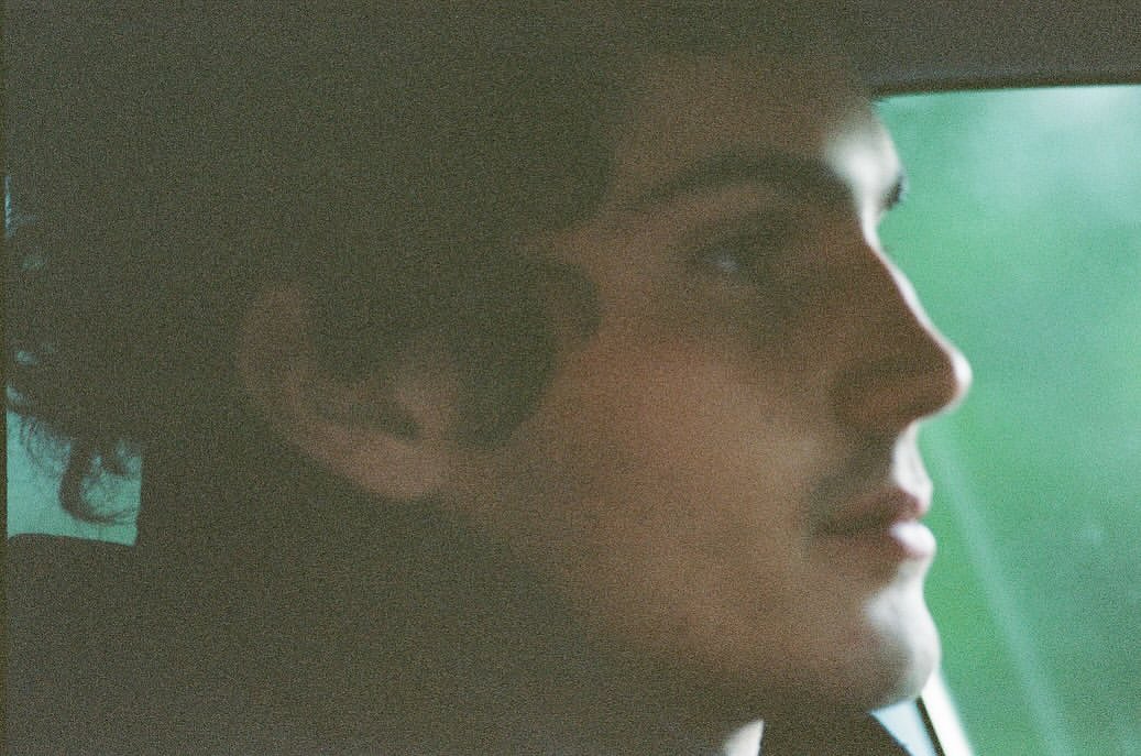 Profile of a young adult male in car looking toward window with natural light, soft focus, and green hues.