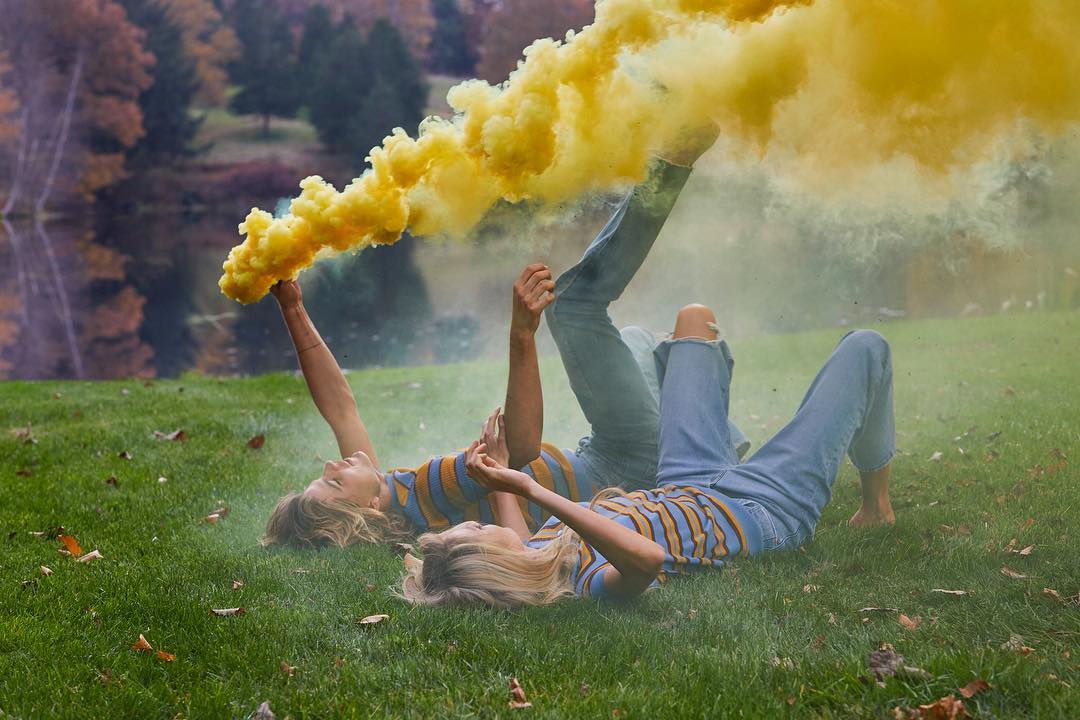 Two adults lying in green grass holding a yellow smoke bomb, with a lake and trees in the background, wearing casual clothes and blue jeans.