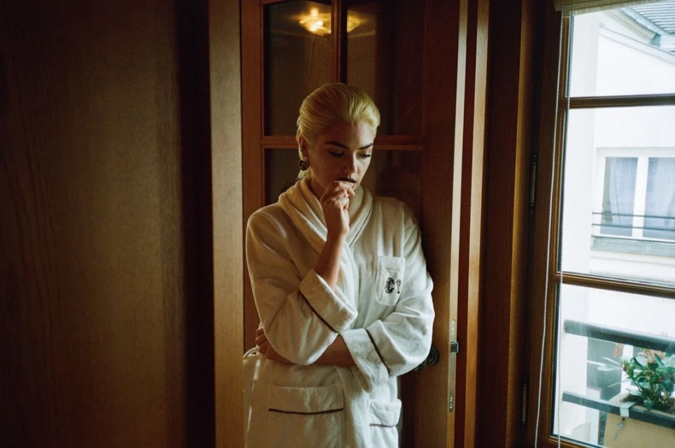 Adult female in a white bathrobe stands in a doorway in an indoor setting, with natural light from a nearby window.