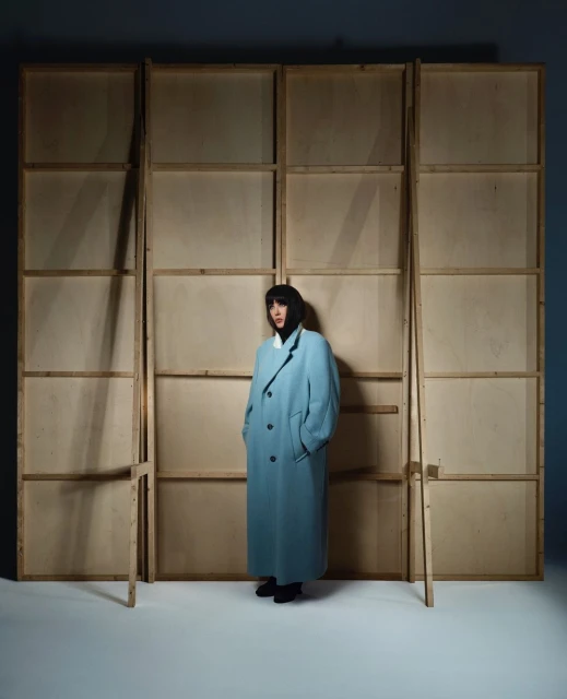 Woman wearing a light blue coat standing in front of a wood frame structure in a photo studio with a neutral background.