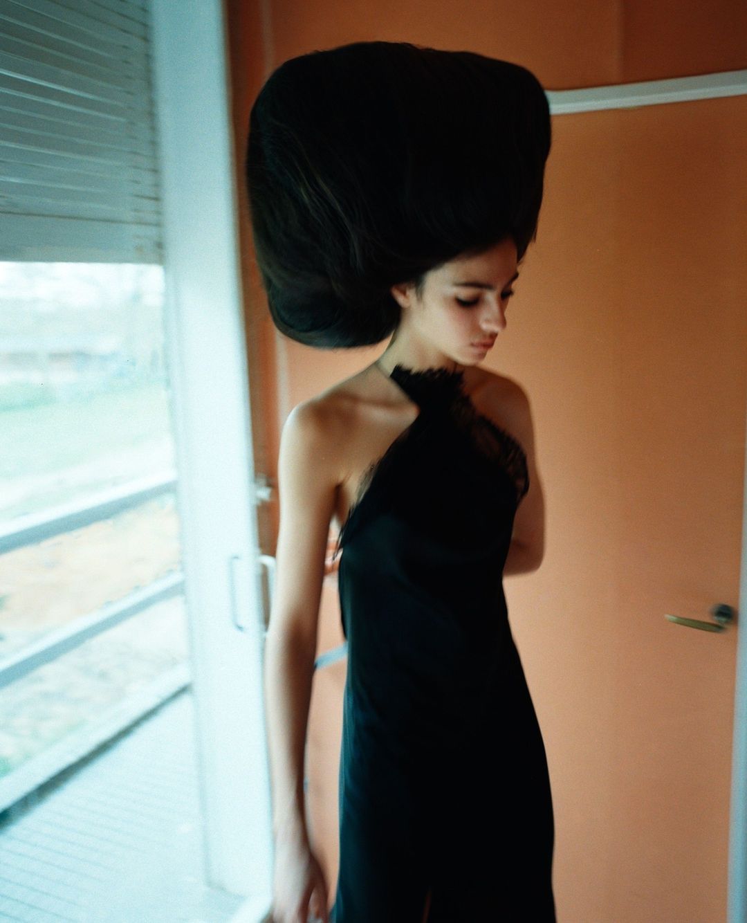 Portrait of a young woman in a black dress with an oversized hairpiece, looking down in an indoor setting, featuring a modern aesthetic.