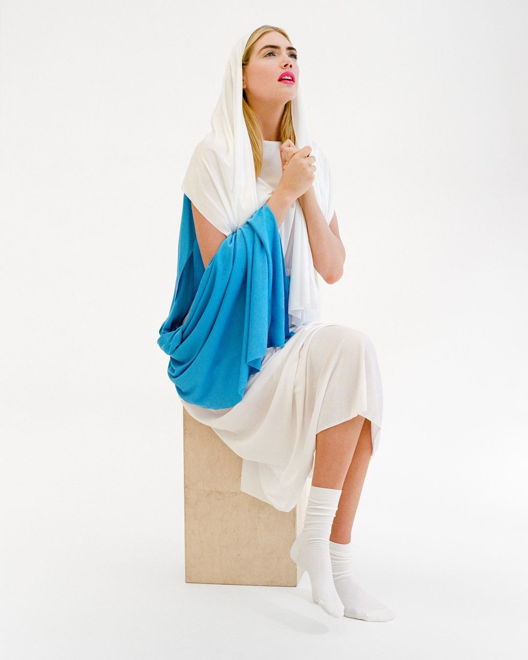 Woman in white dress and headscarf sitting on cube with hands clasped, looking upwards in a brightly lit, neutral indoor setting.
