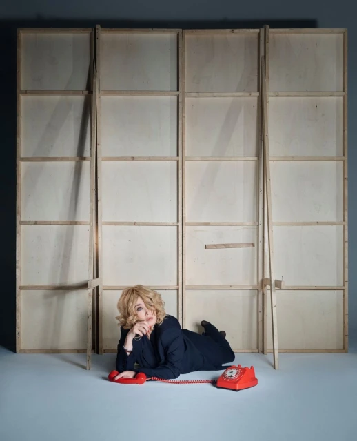 Blonde woman in dark blazer lying on the floor with a red rotary telephone next to a wooden frame structure indoors.
