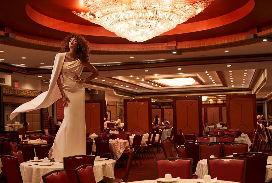 Elegant woman in white dress posing on a table in a banquet hall with a large chandelier and red chairs and tables.