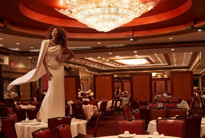 Elegant woman in white dress posing on a table in a banquet hall with a large chandelier and red chairs and tables.