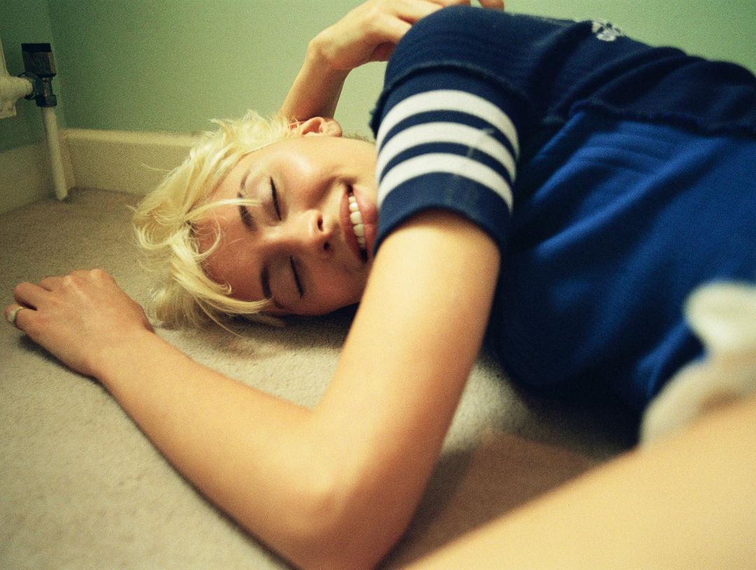 A woman with short blonde hair and a blue shirt lies smiling on the floor in an indoor setting with a neutral background.