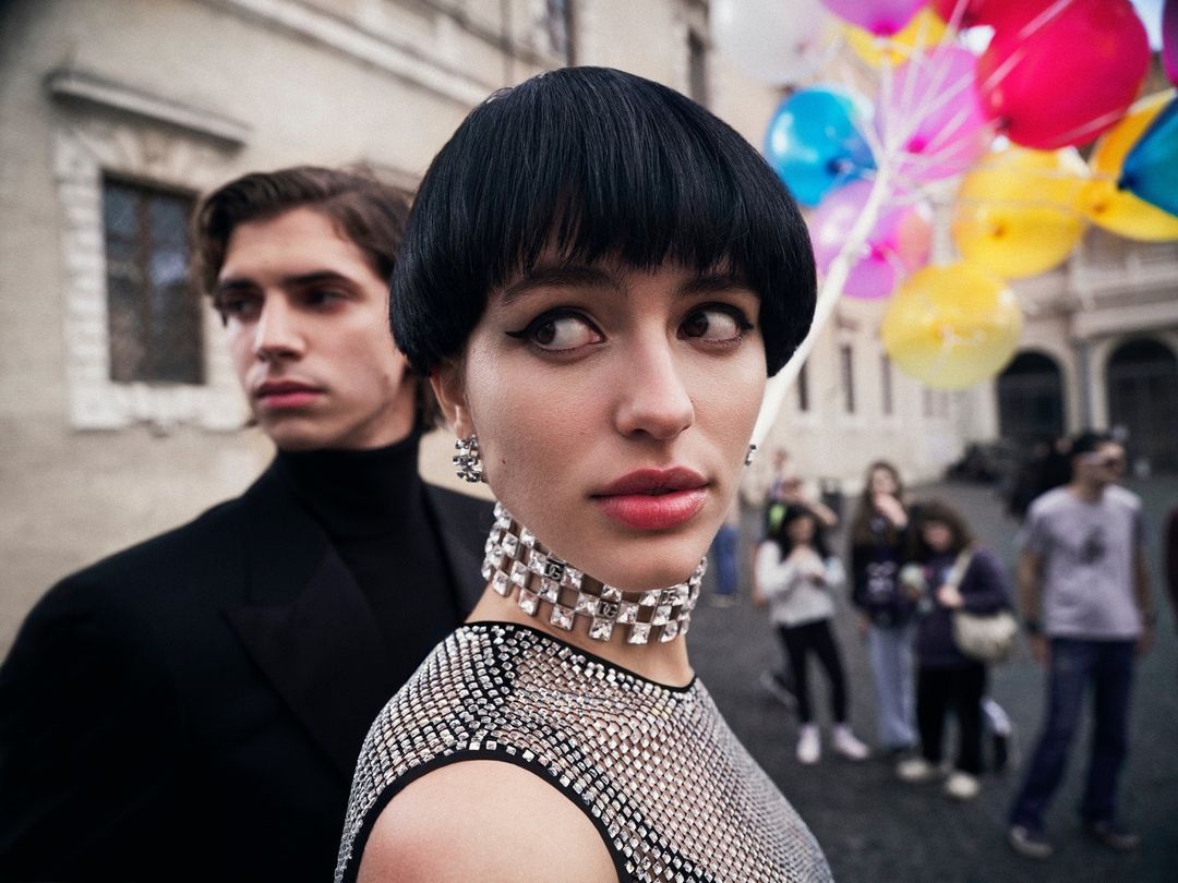 Close-up portrait by Sebastian Faena of a woman with a black bowl cut and crystal choker, with a man standing behind her outdoors.