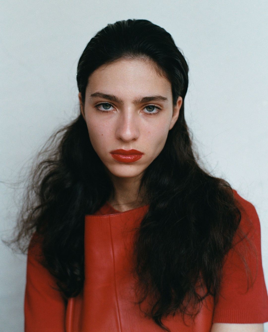 Close-up studio portrait of a young adult woman with dark hair and red lipstick wearing a matching red top against a plain white background.