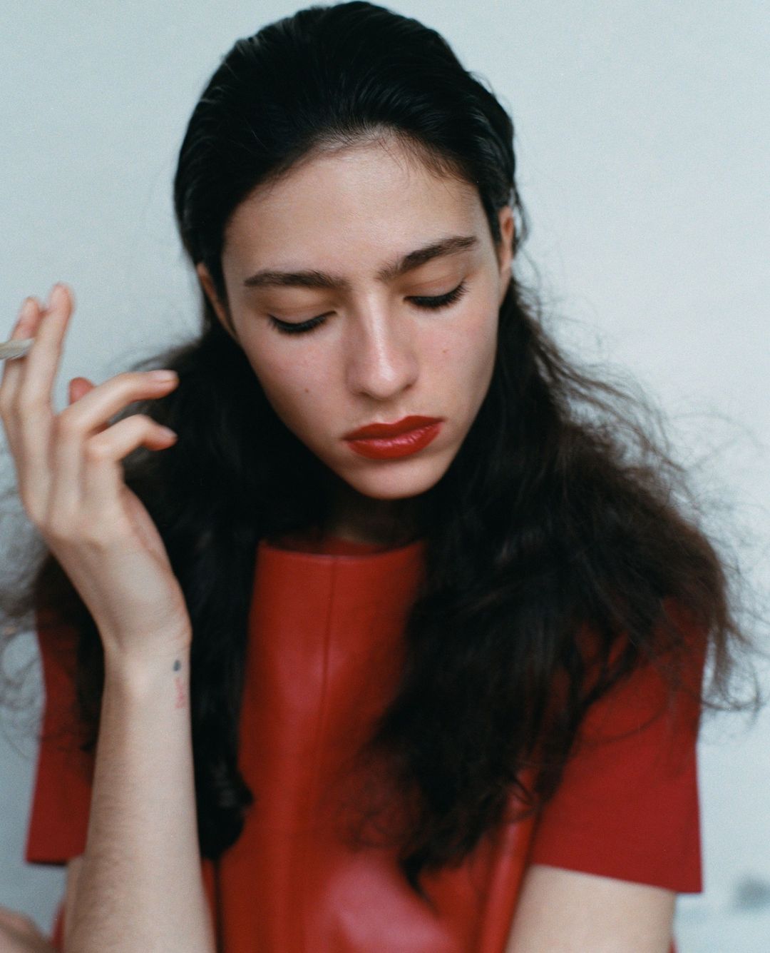 Portrait of a woman with dark hair wearing a red top and holding a cigarette, set against a plain background, fashion or beauty shot.
