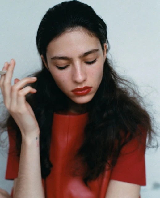 Portrait of a woman with dark hair wearing a red top and holding a cigarette, set against a plain background, fashion or beauty shot.
