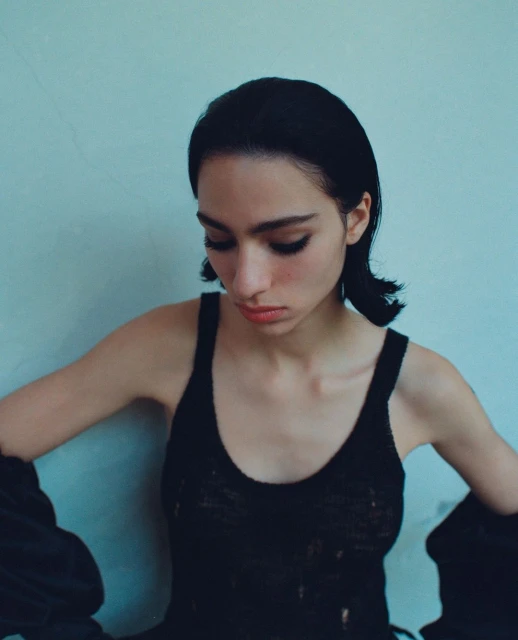 Close-up portrait of a young woman with dark hair wearing a black top against a light blue wall, looking down with a neutral expression.