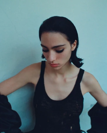 Close-up portrait of a young woman with dark hair wearing a black top against a light blue wall, looking down with a neutral expression.