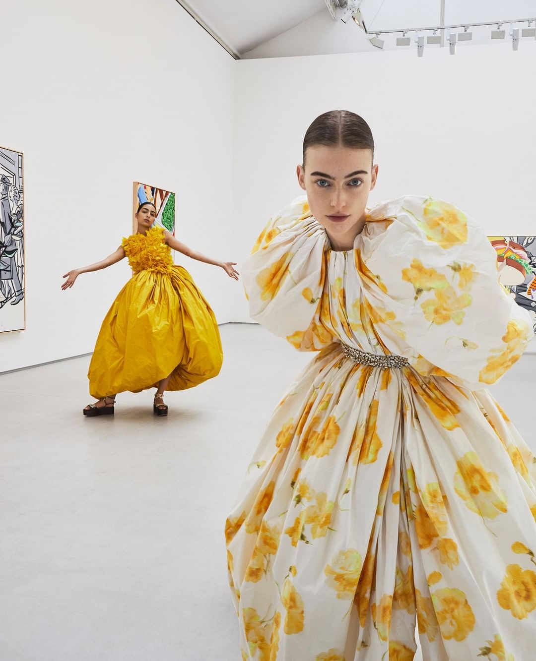 Two women in ballgowns pose in an art gallery. One model in floral pattern stands in the foreground, another in yellow is farther away.
