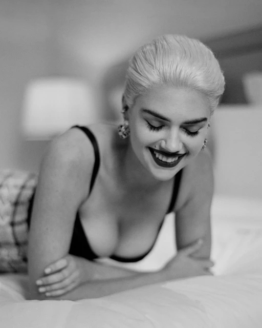 Black and white image of a woman with short blonde hair, lying on a bed, wearing a black bra and looking down with a slight smile.