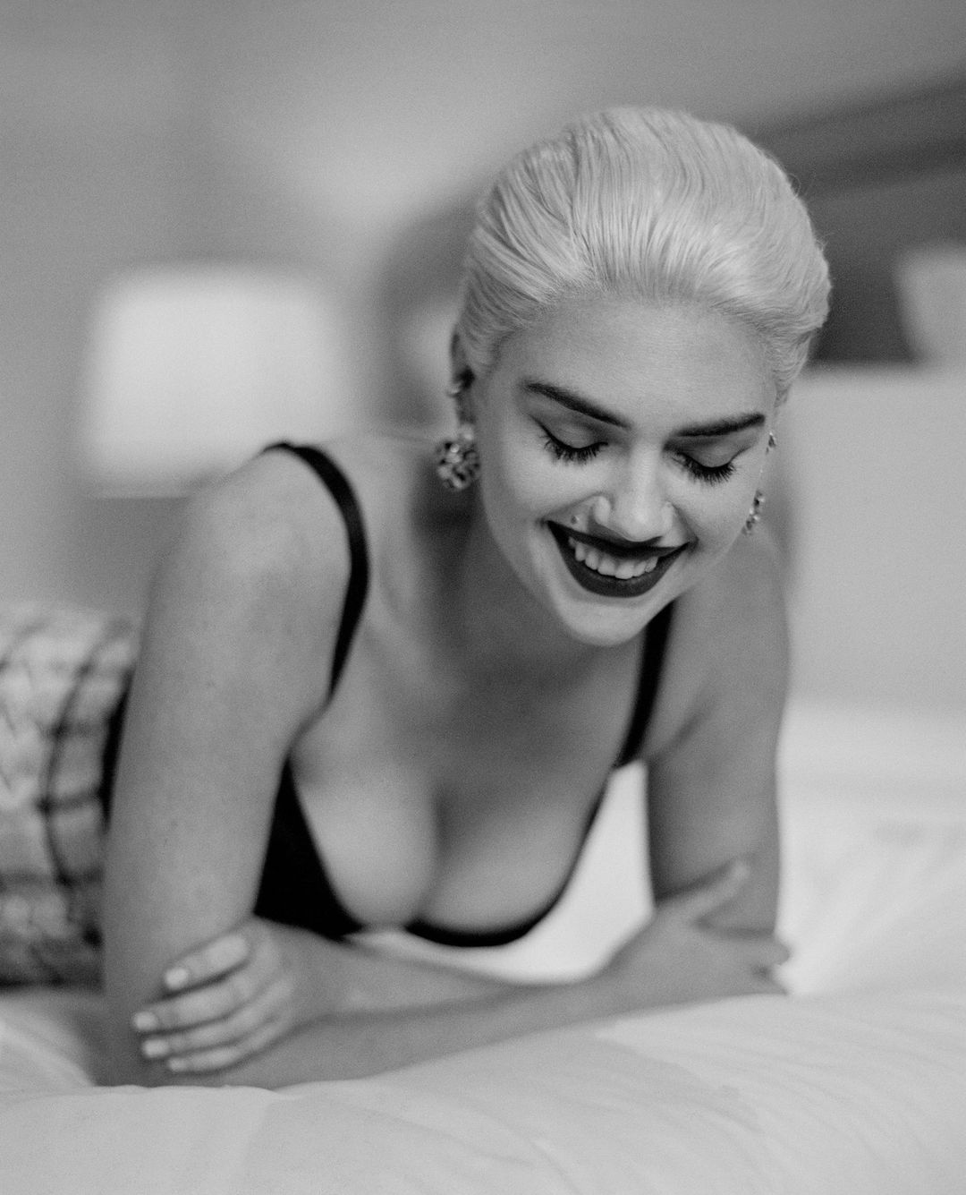 Monochrome portrait of a smiling adult woman with blonde hair lying on a bed in a black bra and patterned shorts, with indoor lighting.