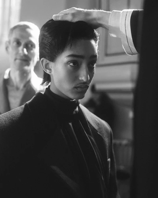 A young adult poses in a monochromatic portrait while a hand rests atop his head, another person stands behind him in a stark black and white image.