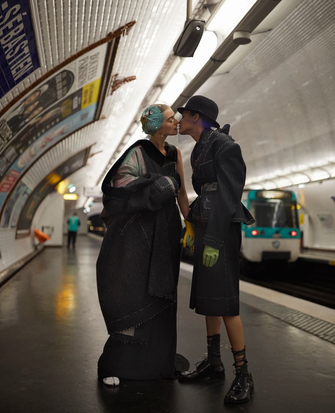 Two people in stylish black outfits share a kiss at a subway station, with a train in the background and subway tiles around them.