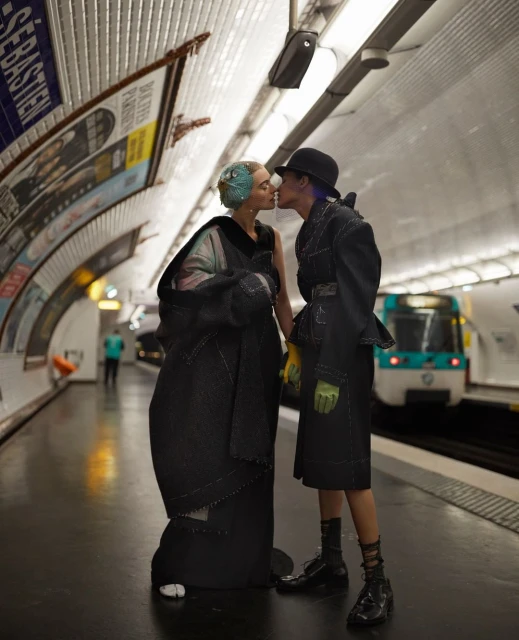 Two people in stylish black outfits share a kiss at a subway station, with a train in the background and subway tiles around them.