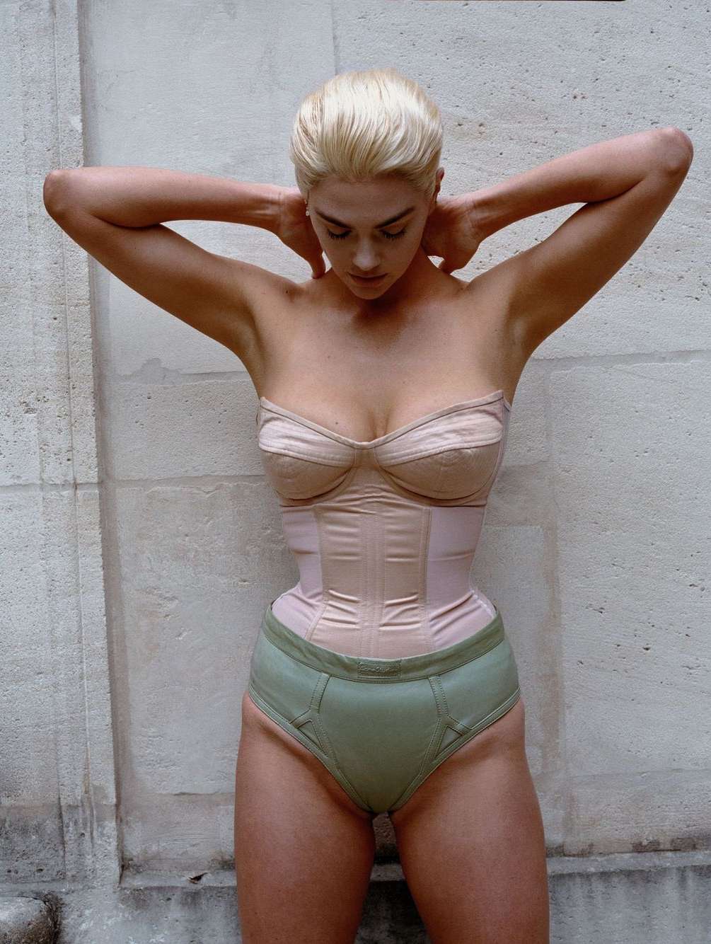 Woman with platinum blonde hair poses in a tan corset and underwear in front of a weathered white wall in a full shot.