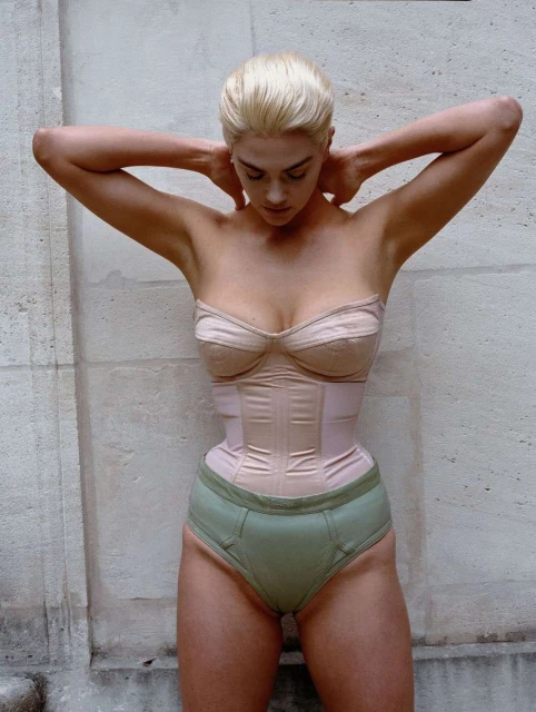 Woman with platinum blonde hair poses in a tan corset and underwear in front of a weathered white wall in a full shot.