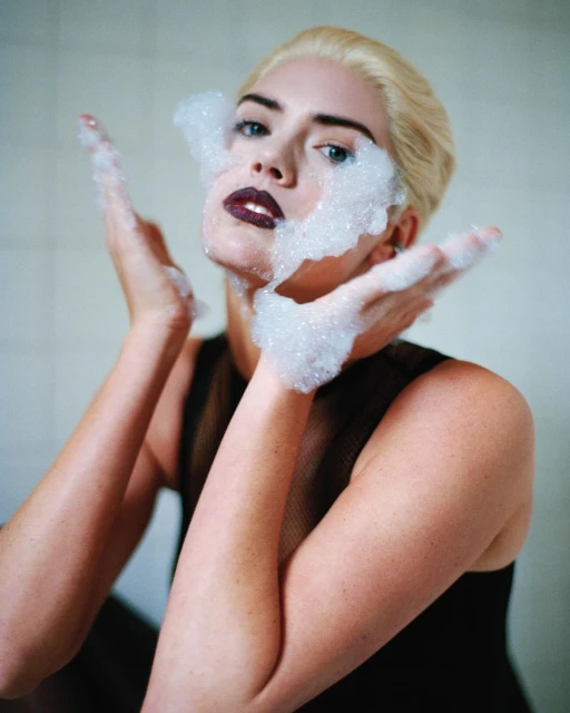 Portrait of a fair-skinned woman with blonde hair and dark lipstick, face partially covered in white soap bubbles, holding her hands up.