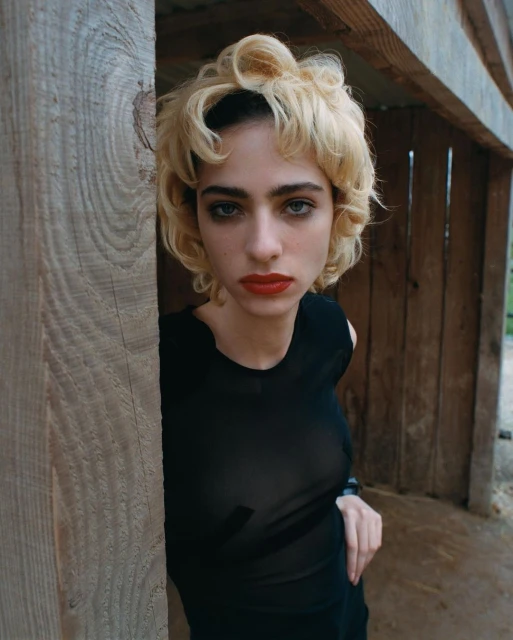 Blonde woman with red lipstick poses in front of a wooden structure, wearing a black top and looking directly at the camera.