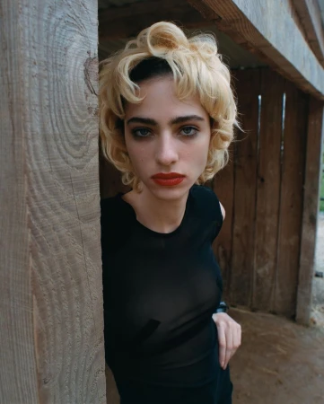 Portrait of a woman with short blonde curly hair and red lipstick wearing a black sleeveless top leaning against a wooden post