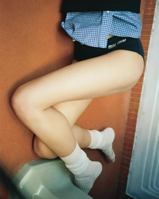 Woman in black briefs and white socks lies on a terracotta tile floor, wearing a blue and white gingham shirt.