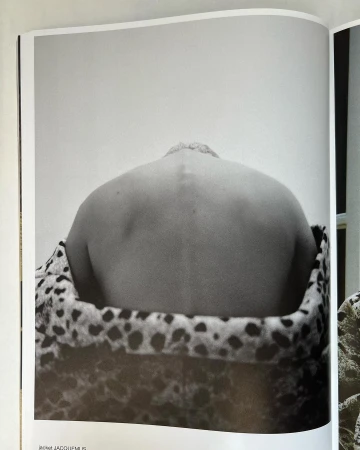 Black and white photo of a person's back and shoulders against a plain wall, draped with a patterned animal-print fabric.
