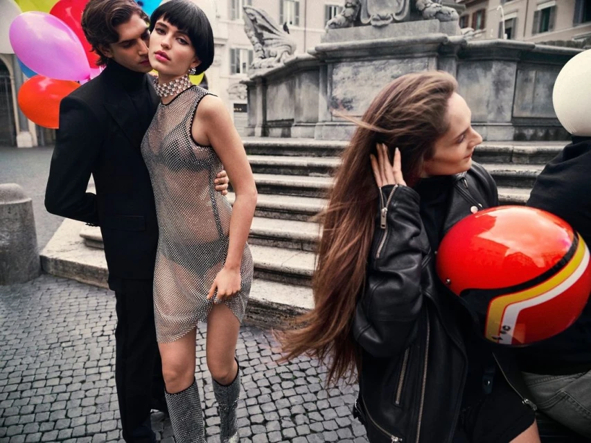 A stylish couple poses on a cobblestone street with balloons, while a woman in a leather jacket sits on a motorcycle nearby a stone fountain.