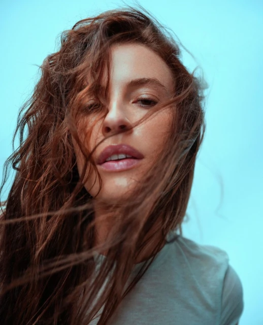 Close-up portrait of a young adult woman with auburn hair. She wears a casual grey top against a clear sky backdrop.