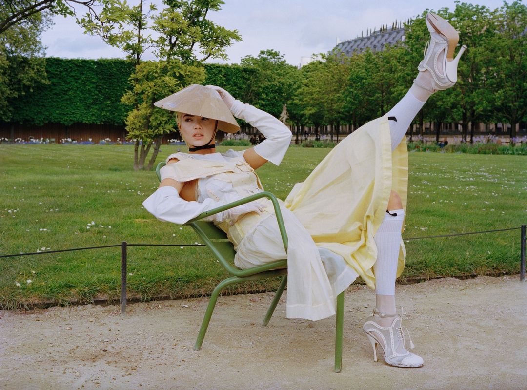 Woman in avant-garde fashion poses on green chair in park, wearing pale yellow dress, wide-brimmed hat, and high-heeled shoes outdoors.