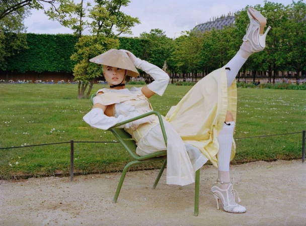 Woman in avant-garde fashion poses on green chair in park, wearing pale yellow dress, wide-brimmed hat, and high-heeled shoes outdoors.
