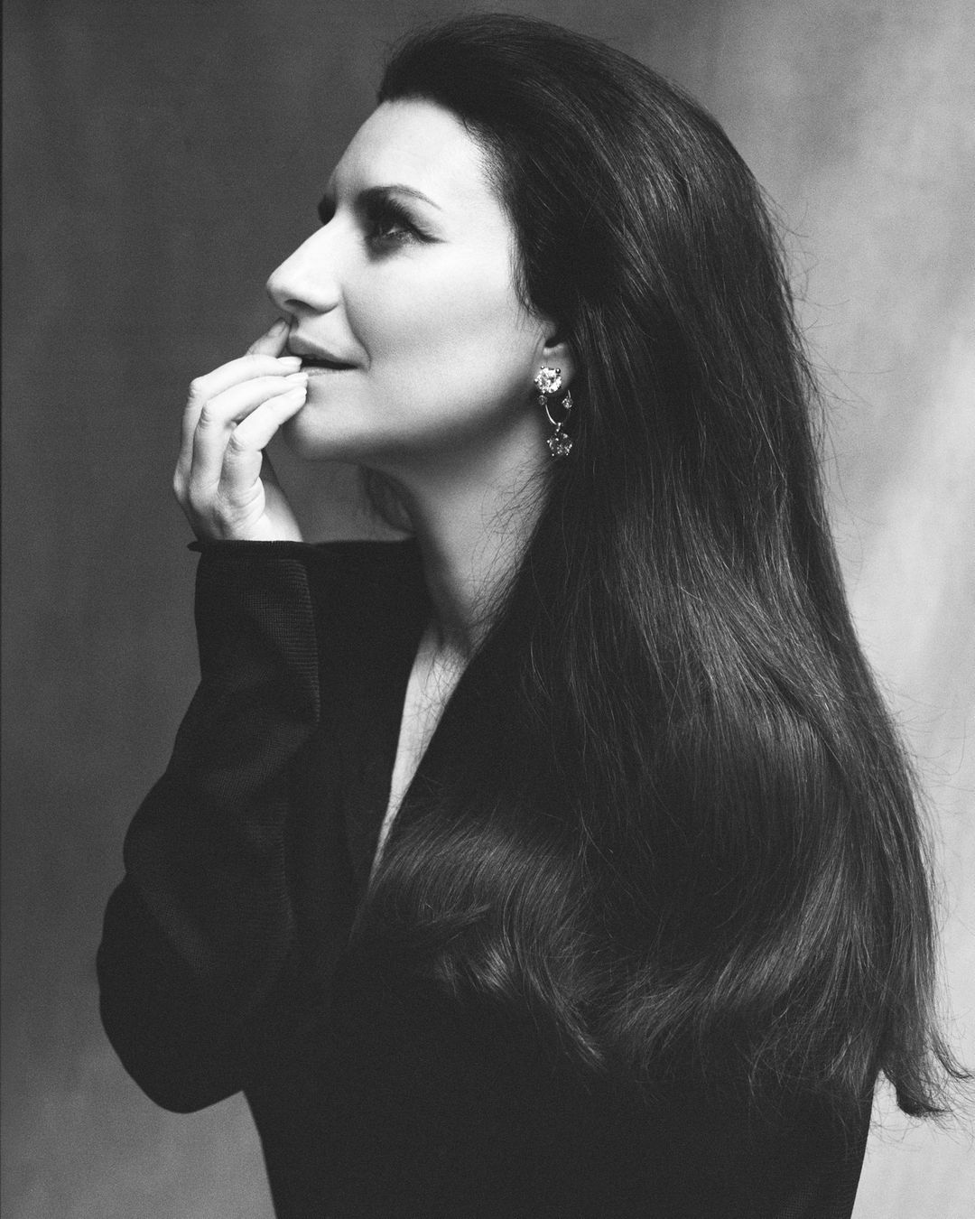 Black and white portrait of a woman with long dark hair in a jacket looking up with hand near face and jewelry earrings.